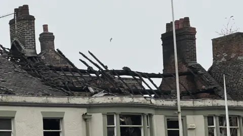 The roof of a building which appears to have been damaged by fire. Chimney pots on either side of the roof appear to be undamaged, but the tiles have been destroyed or removed and only the framework of the roof remains.