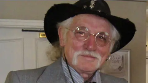 Park dressed in a formal, vintage‑style outfit. He is wearing a light grey three‑piece suit consisting of a jacket, waistcoat, and trousers, paired with a striped collared shirt and a dark patterned tie. A bolo tie cord is also visible around the neck area. He is wearing a wide‑brimmed black hat decorated with subtle embellishments, and he has long, light-colored hair extending past the hat’s brim.