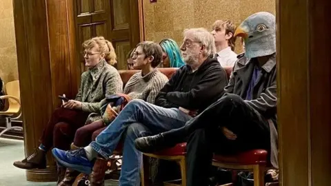 Owen Sennitt/LDRS A row of four people seated on red chairs. The person on the immediate right of the image is wearing black and has his legs crossed. On his head he is wearing a rubber hood-mask of a pigeon's head, obscuring his entire head and face.