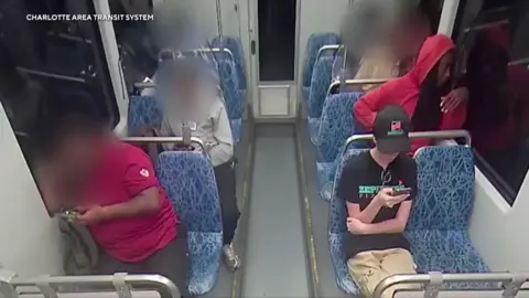 Charlotte Area Transit System A CCTV camera shows a woman, in a black baseball cap, hiding her face, a T-shirt and slacks, on her phone in a train with a man in a red hooded sweatshirt seated behind her. Other people on the bus are blurred out. 