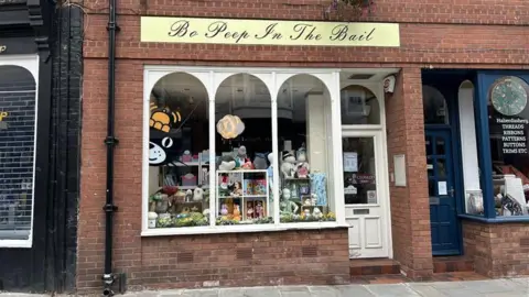 The outside of a shop window with three arched windows and a white door to the right. A black cat with an orange hat logo is peeking out from the left and there are several teddies on shelves in the window on display. At the top of the windows, a yellow and black sign reads "Bo Peep In The Bail".