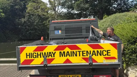 A man is putting a black bag of dead fish into the back of a Highway Maintenance van.