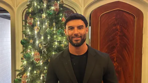Liam Reardon stands in front of a Christmas tree in a dark suit with black T-shirt.