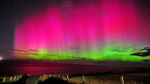 BBC Weather Watchers/Saf37y A picture of the night sky across a big empty expanse. The sky is lit up bright green and pink in waves, showing the aurora, in front of a sparkling night sky filled with stars