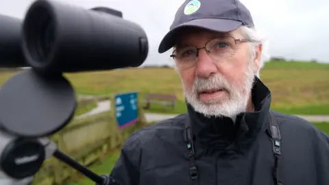 Close-up of a man in a black waterproof jacket and cap with a circular blue RSPB logo, standing outdoors near grassy fields. A large spotting scope on a tripod is positioned in the foreground, angled toward the landscape. A wooden fence and a blue information sign are visible in the background under an overcast sky. He wears glasses and has grey hair under the cap and a grey beard.