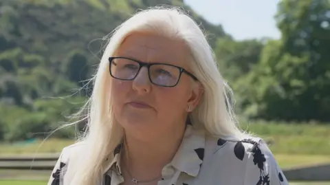 Allison Graham has long blonde hair and wears a black and white patterned shirt and dark glasses. She is sitting outside and there is greenery behind her 
