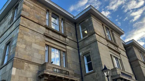 Light coloured brickwork at the back of the building. A huge stately home with long windows and stone balconies.