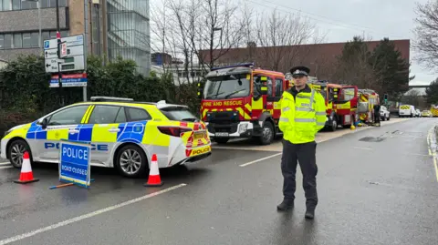 A police officer stands next to a police car at a road block. Behind him are three fire engines parked by the side of the road. 