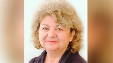 A woman with short curly blonde hair is pictured smiling while wearing a brown cardigan. 