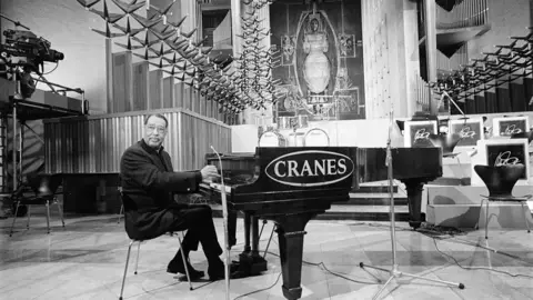Getty Images A black and white image of Duke Ellington in Coventry Cathedral. He is sitting at a grand piano in front of the cathedral's altar and famous Graham Sutherland tapestry