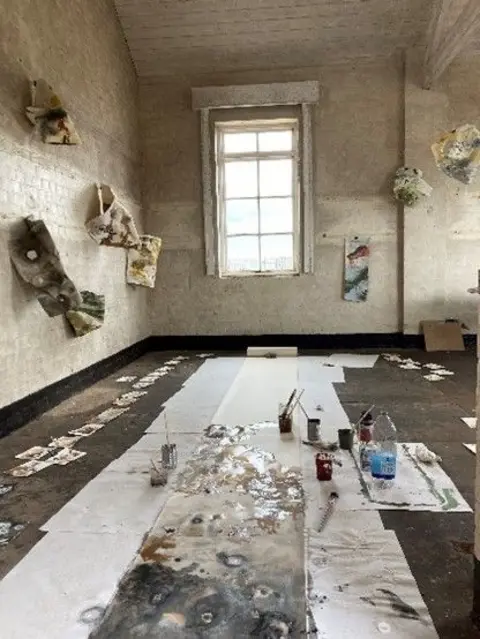Jane Watt The inside of a temporary art studio on Orford Ness. On the walls are two and three-dimensional paintings. The floor is covered in white pieces of paper and paint brushes and paint pots.