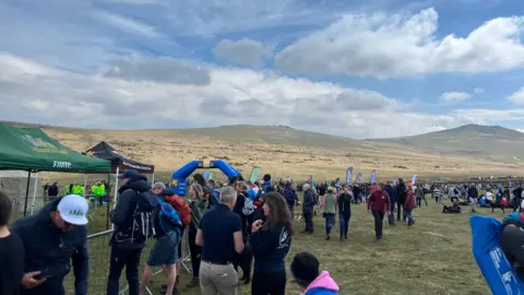 There are hundreds of people lining the fencing waiting to welcome the walkers. Dartmoor is lit with sunshine.