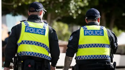 Two police officers on patrol, facing away from the camera.Both are wearing blue shirts and yellow police jackets. The background is blurred.