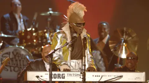 Getty Images Sly Stone is pictured later in his life wearing a huge white leather belt with the word 'Sly' embellished in silver. He has a matching silver jacket with the collar popped, and a tall bleached mohawk. He plays a white keyboard and sings into two microphones on stands.