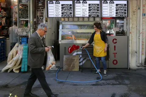 Getty Images A man walks by and a woman cleans the pavements with a hose outside a small restaurant in Tehran