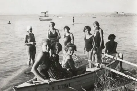 Church of Scotland Miss Haining is in the background up to her knees in water. She is surrounded by girls - five of whom are in a rowing boat in the foreground. It is a black and white picture.