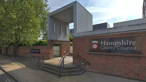 Google Google street view image of hampshire cc's office. It has two red brick walls with the councils name on signage. There is a gap between the walls which is covered by a silver structure.