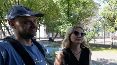 Andrei and his wife Yekaterina are pictured talking to the BBC as cars can be seen queuing in the background. 