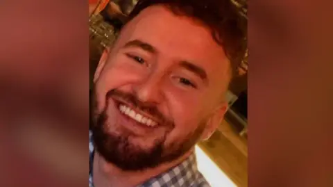 A smiling Conor Browne is wearing a checked shirt. He has short hair and a well groomed beard.