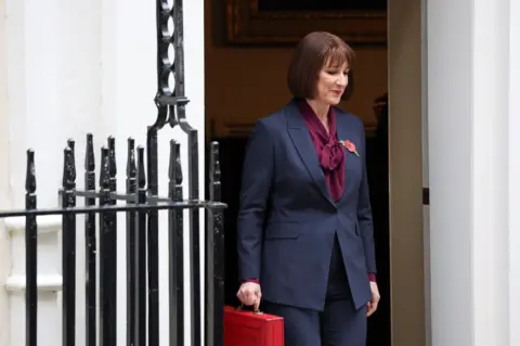 PA Media Rachel Reeves comes through the doors of 11 Downing Street holding her red budget box and wearing a blue suit, burgundy top and red poppy. There is a black railing in front of the white building.