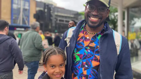 Machel with his daughter, both smiling into the camera. Machel is wearing a brightly coloured shirt with a blue jacket, as well as a baseball cap. 
