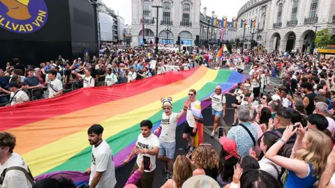 Getty Images A large crowd of spectators lines the street as a group of parade participants carries a massive, elongated rainbow Pride flag down a central London road flanked by historic stone buildings.