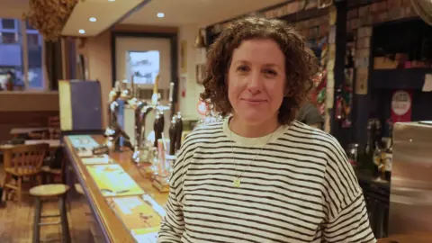 Cerys Furlong, with curly hair and a striped top, stands at the bar in The Grange pub in Grangetown, Cardiff. A row of beer pumps in soft focus behind Cerys, with warm lighting.