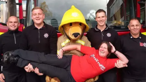 Surrey Fire and Rescue Service A member of the charity lying in firefighters arms in front of a fire engine