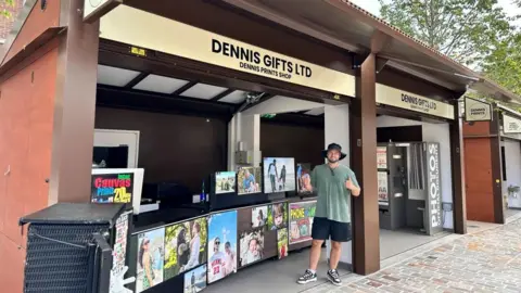 Carrol Weston/BBC Man in green hat and T-shirt standing with thumbs up in front of a wood-coloured stall selling paintings. Various pieces of artwork are on display.