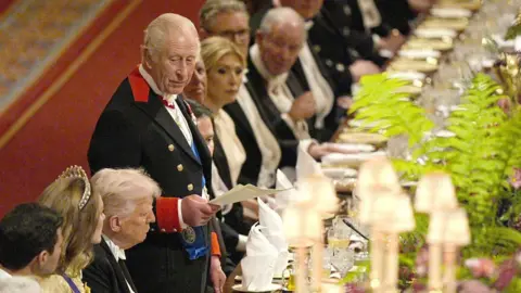 King Charles stands at a large banquet table and appears to be delivering a speech from a piece of paper. The US president Donald Trump is sat in a chair next to him. 