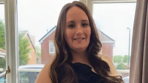 A woman in her early 30s with long, wavy brown hair and wearing a black sleeveless dress, stands in front of a window smiling at the camera. 