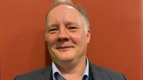 A man with short grey hair, a pale shirt and a grey jacket in front of an orange background