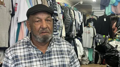 A man looking straight at the camera with a neutral expression. He is wearing a black cap and a black and white checked shirt and is standing in a clothes shop in front of a rail of T-shirts.