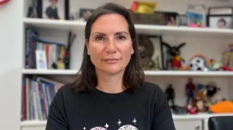 Natalie Neri looks into camera with photos of her son, books, and toys on the shelf in the background. She has shoulder-length dark hair and is wearing a black t-shirt with white stars on