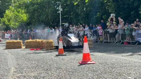 A person wearing all black riding a black kart between straw bales and orange cones over a ramp. Crowds of people are watching from behind metal railings. 