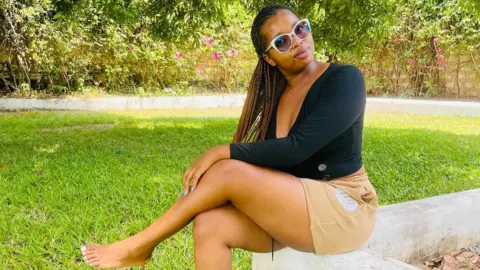 Naomi Kuria/Facebook Naomi Kuria in a black top and brown shorts, sitting in a compound with green grass