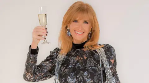 BBC Jackie Bird smiles at the camera while raising a glass of wine. She has strawberry blonde hair and wears a sparkly silver and black long-sleeved top.