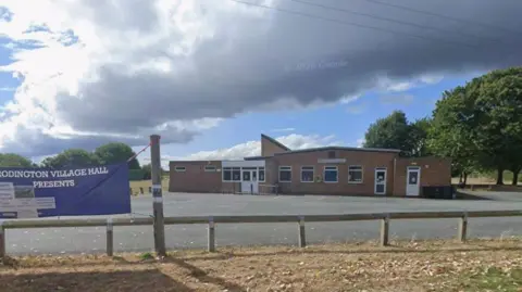 A building with a forecourt, viewed from across a roadside fence under a cloudy sky.