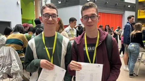 Adrian and Łukasz Koman, two identical twins, smile while each holding a sheet of paper detailing their A-level results. Both boys wear black glasses, have short brown hair, and are wearing bright yellow lanyards. They are stood in a busy school hall with pupils and staff milling about in the background.