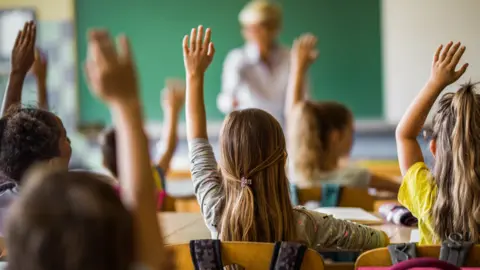 A classroom of children sitting at desks and holding their hands in their air to answer a teachers standing in front of a board at the front of the class.