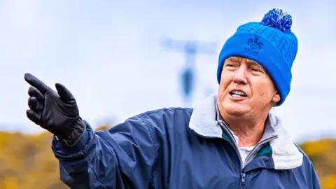 Donald Trump wearing a blue jacket and blue bobble hat on the site of his new course near Menie, Aberdeenshire