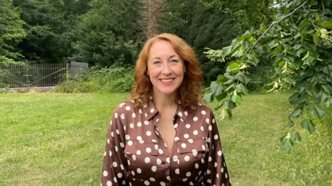BBC/Sofía Luis-Hobbs A woman with auburn hair smiles at the camera in a green open wooded area. She wears a brown button down dress with white polka dots.