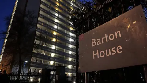 A picture of Barton House taken at night. It shows the outdoor corridors of the tall tower block lit up in the darkness, and signage outside the front gate.