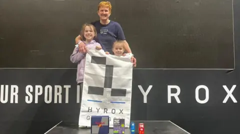 Stuart Amory Carole Munro and her two granddaughters aged eight and four standing on a podium holding a white and black Hyrox flag. 