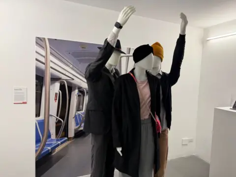 A plain white room with three dummies. One is dress as a woman and two as men. They are posed like they are posed as if they are riding the underground or metro with a display of a carriage behind them. 