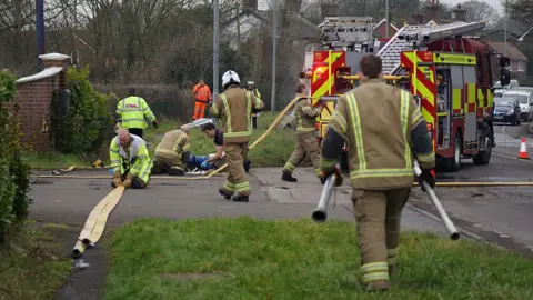 Dan Jessup Fire crews and a fire engine block a road with a grassy area to the side. Crews are preparing a fire hose, some knelt on the tarmac, some walking. To the right, a line of cars is visible, heading in the opposite direction.