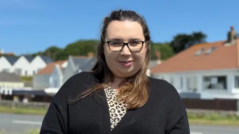 A women with chest length brown hair. She is looking at the camera and smiling. She has glasses on. She is wearing a black thin cardigan and a leopard print top underneath.