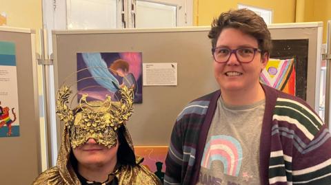Two people, one wearing a golden floral mask and hooded cloak and the other in a cardigan, grey tshirt and wearing glasses