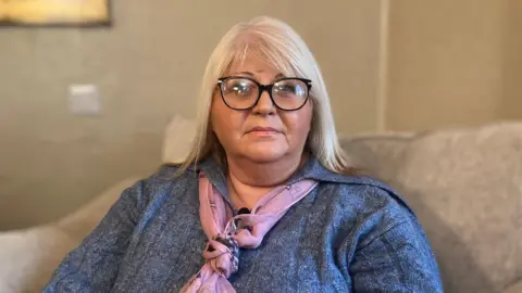 BBC A woman with blonde hair, wearing glasses. She is sat on a a light-coloured sofa. She is wearing a blue top with a pink scarf.