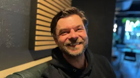 A man called Alex Morrall smiles while sat inside a bar run by Verdant Brewery in Cornwall. Mr Morrall is wearing a black hoodie and has a beard.